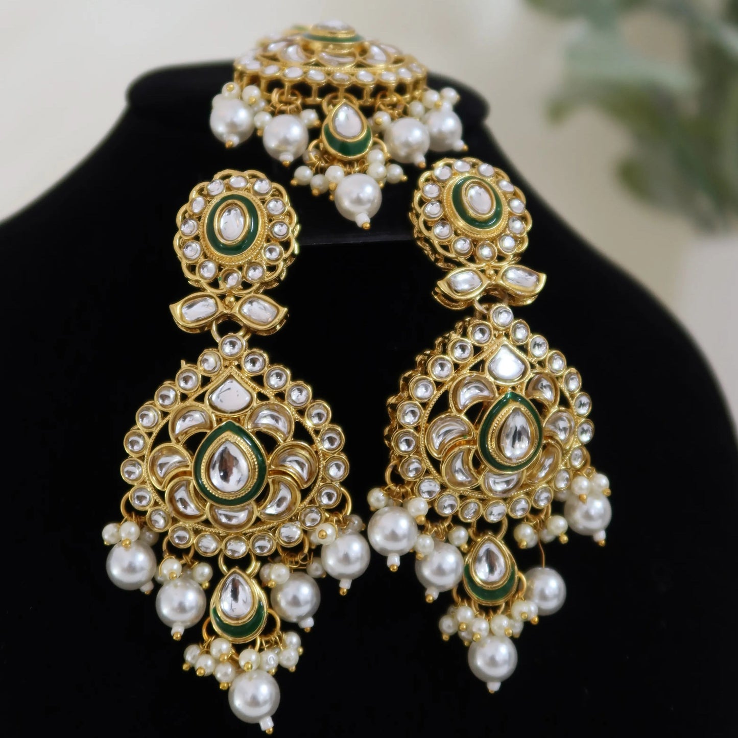 Ornate gold and pearl earrings on a black stand with a blurred plant in the background