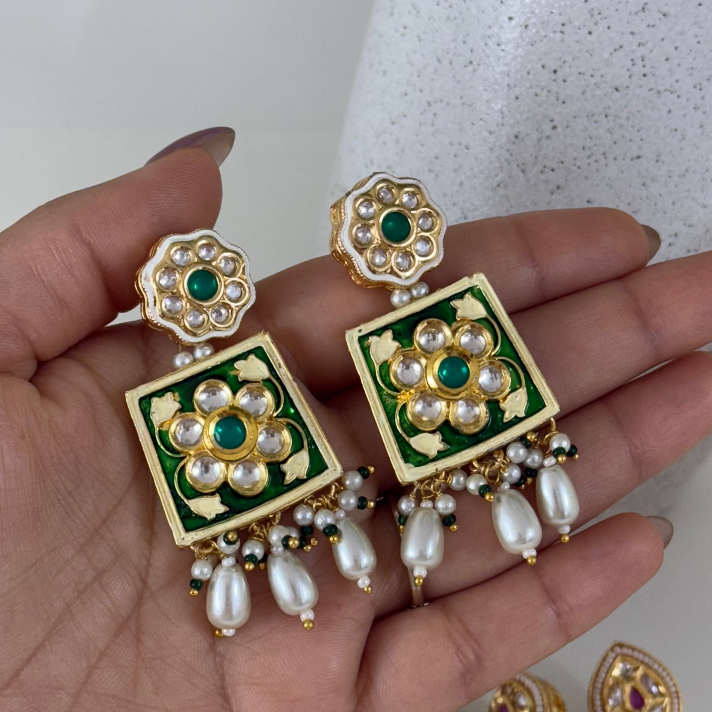 Ornate earrings with green and gold design held in a hand against a neutral background
