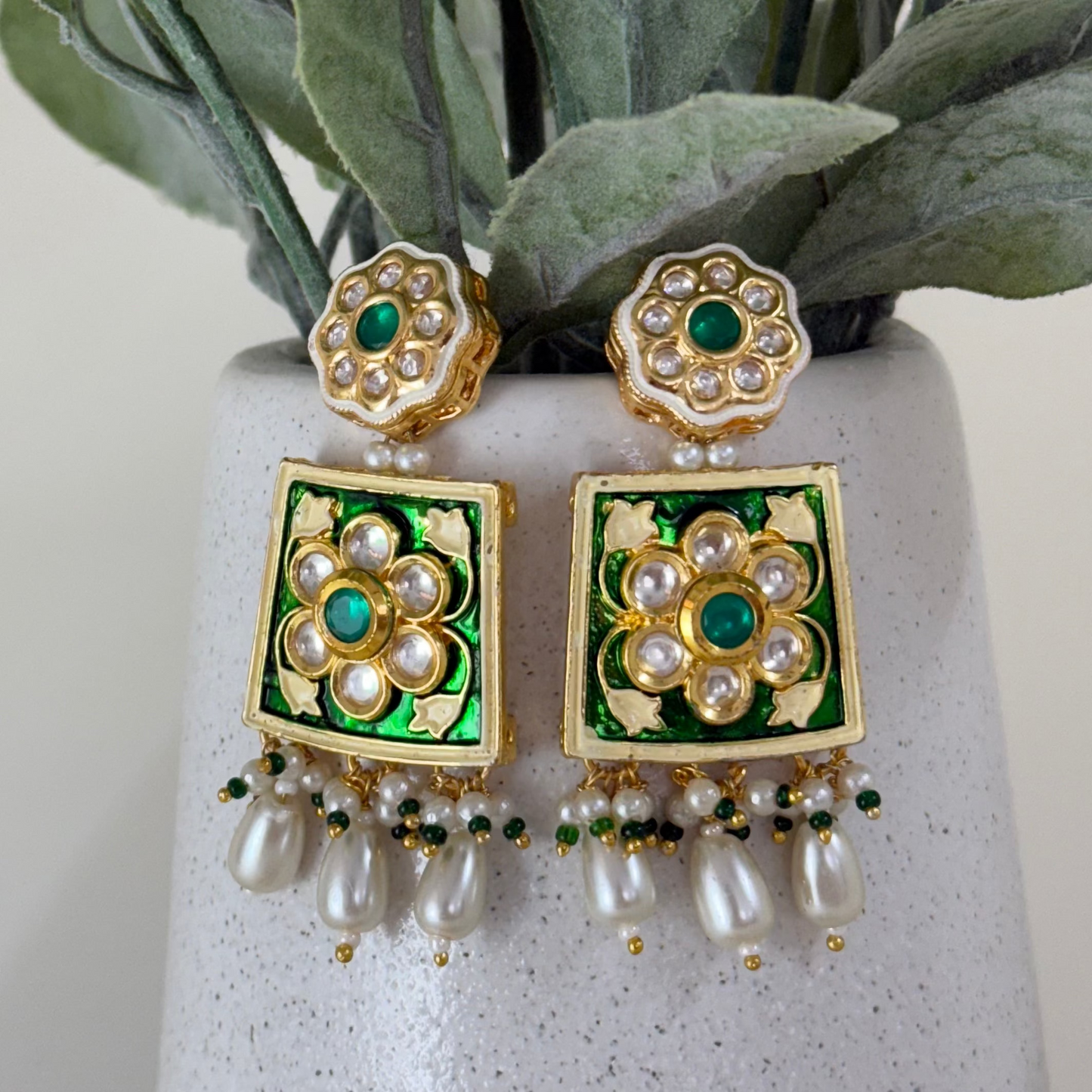 Decorative earrings with green and gold accents on a white vase with green leaves.