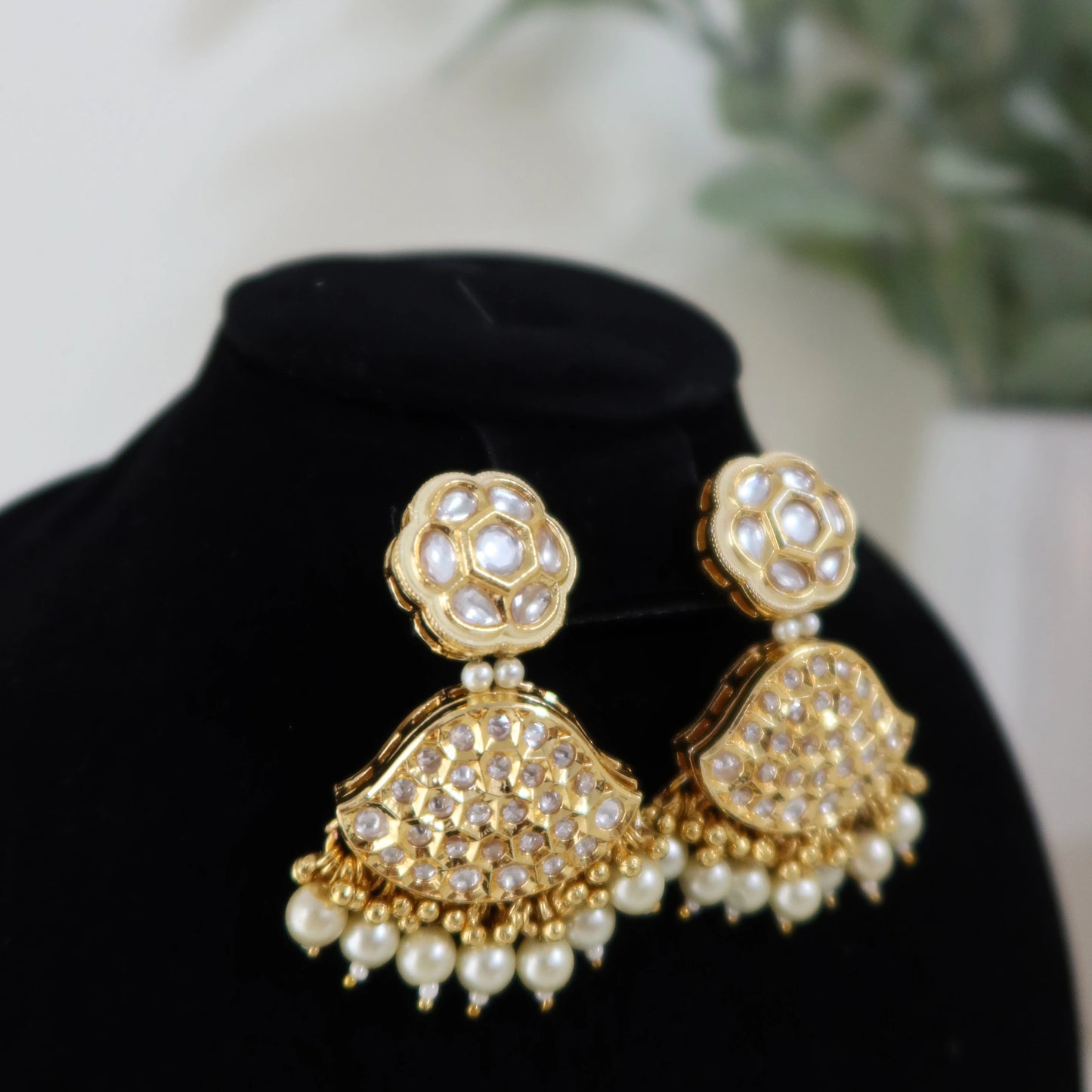 Gold and pearl earrings on a black stand with a blurred green plant background