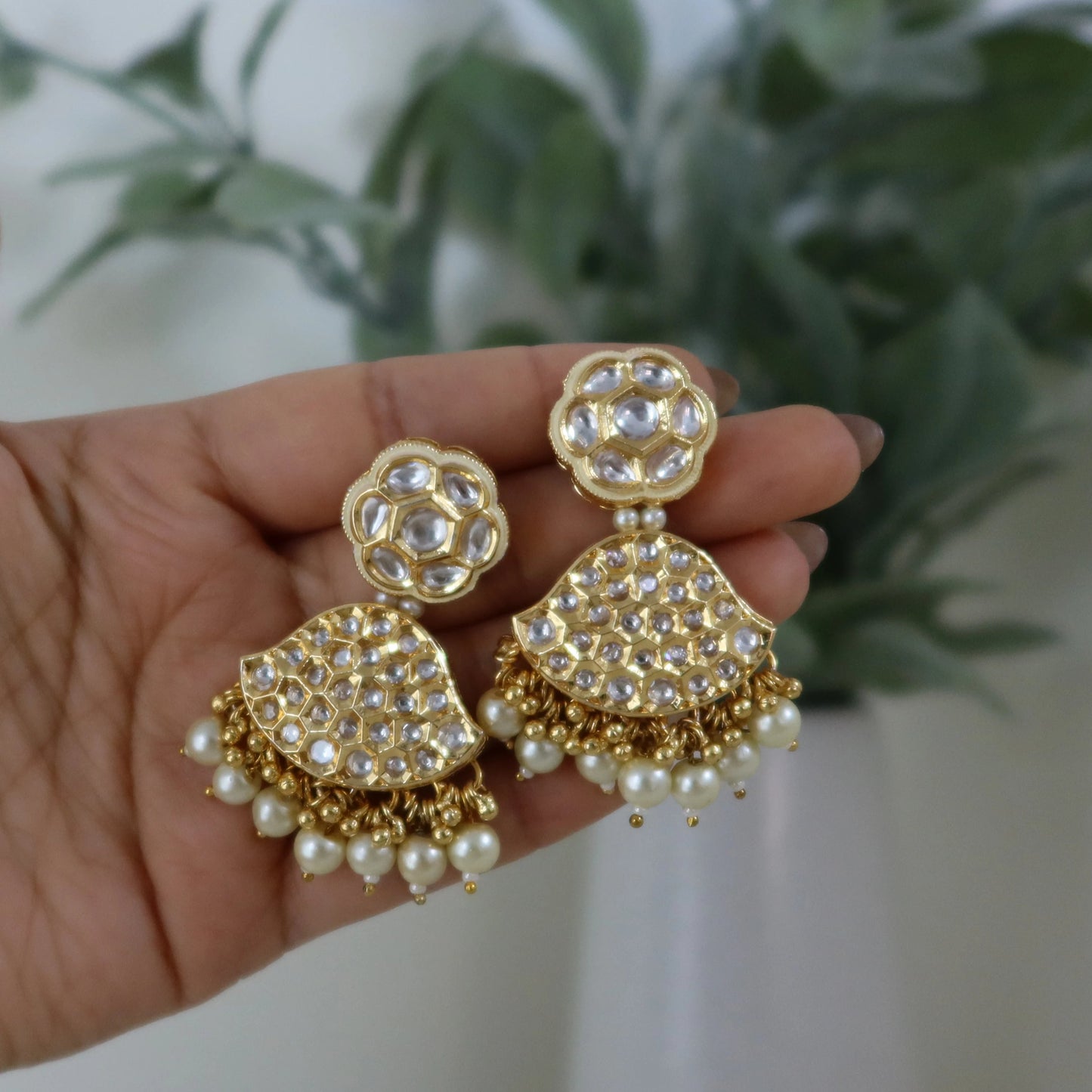 Gold and pearl earrings held in a hand with a blurred plant background