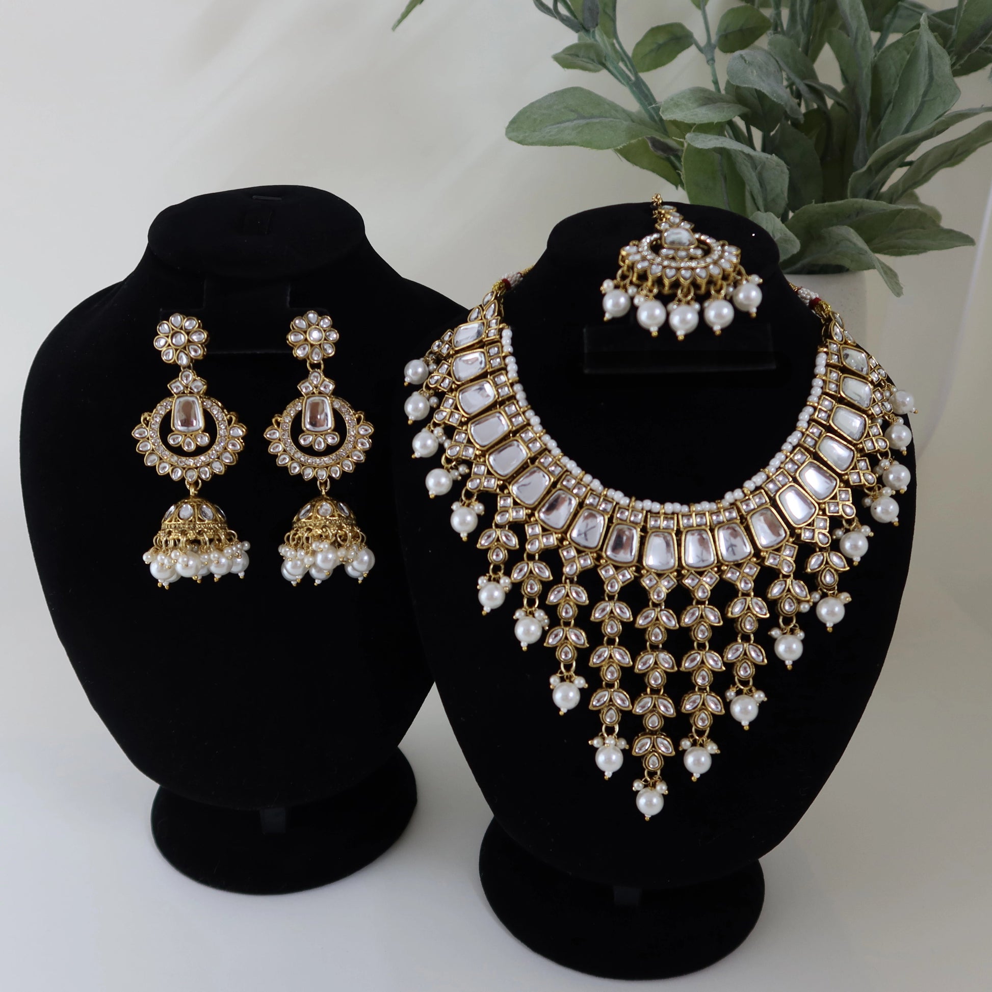 Gold and pearl necklace and earrings set on black stands with a plant in the background.