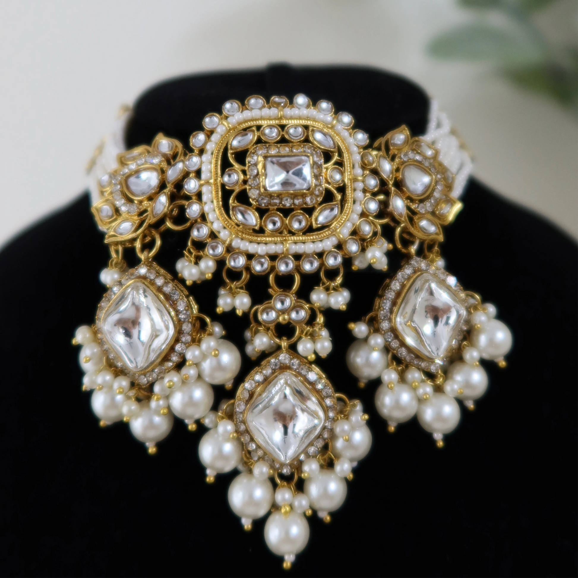 Ornate gold and pearl earrings on a black stand with a blurred green plant in the background