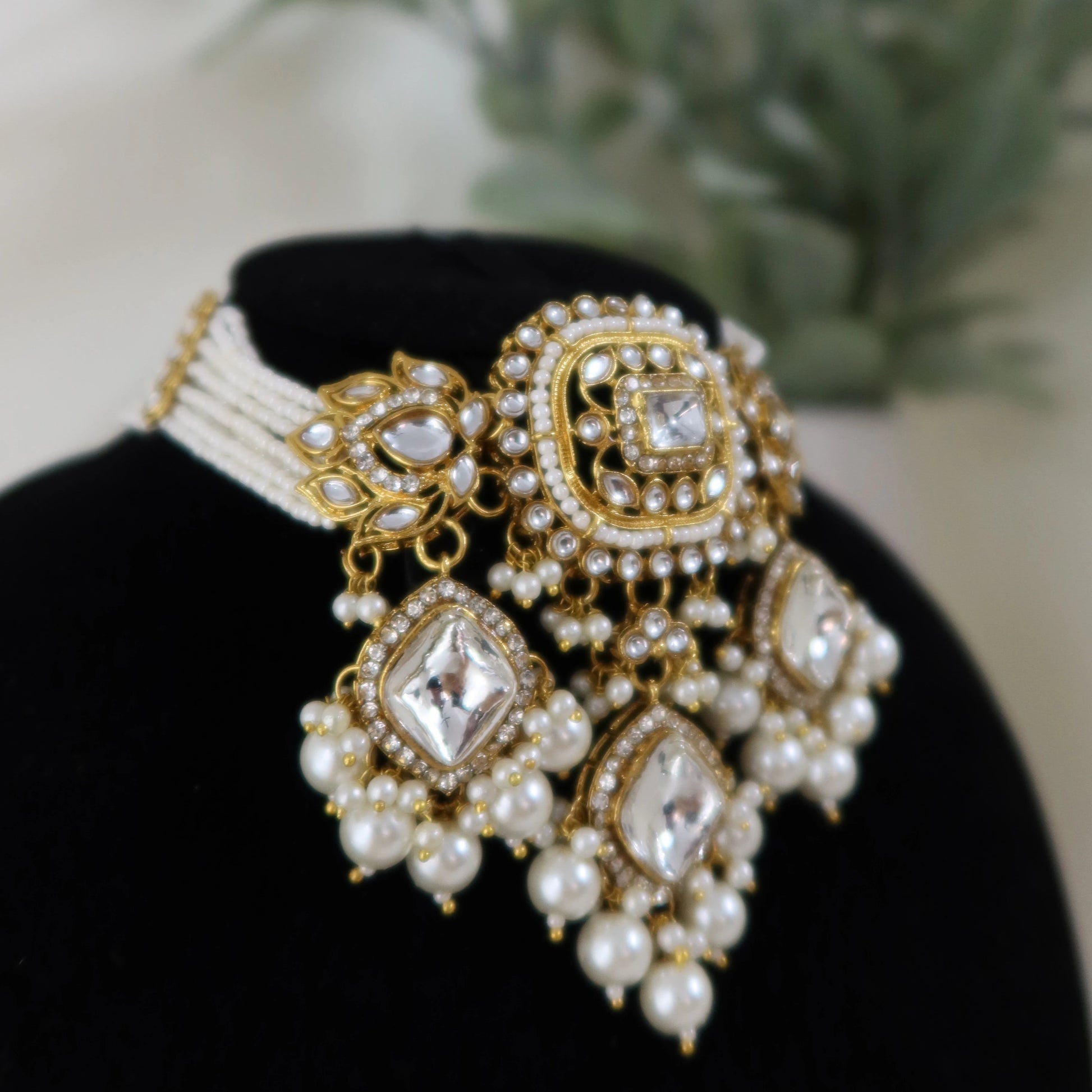 Ornate gold and pearl necklace on a black stand with a blurred plant in the background
