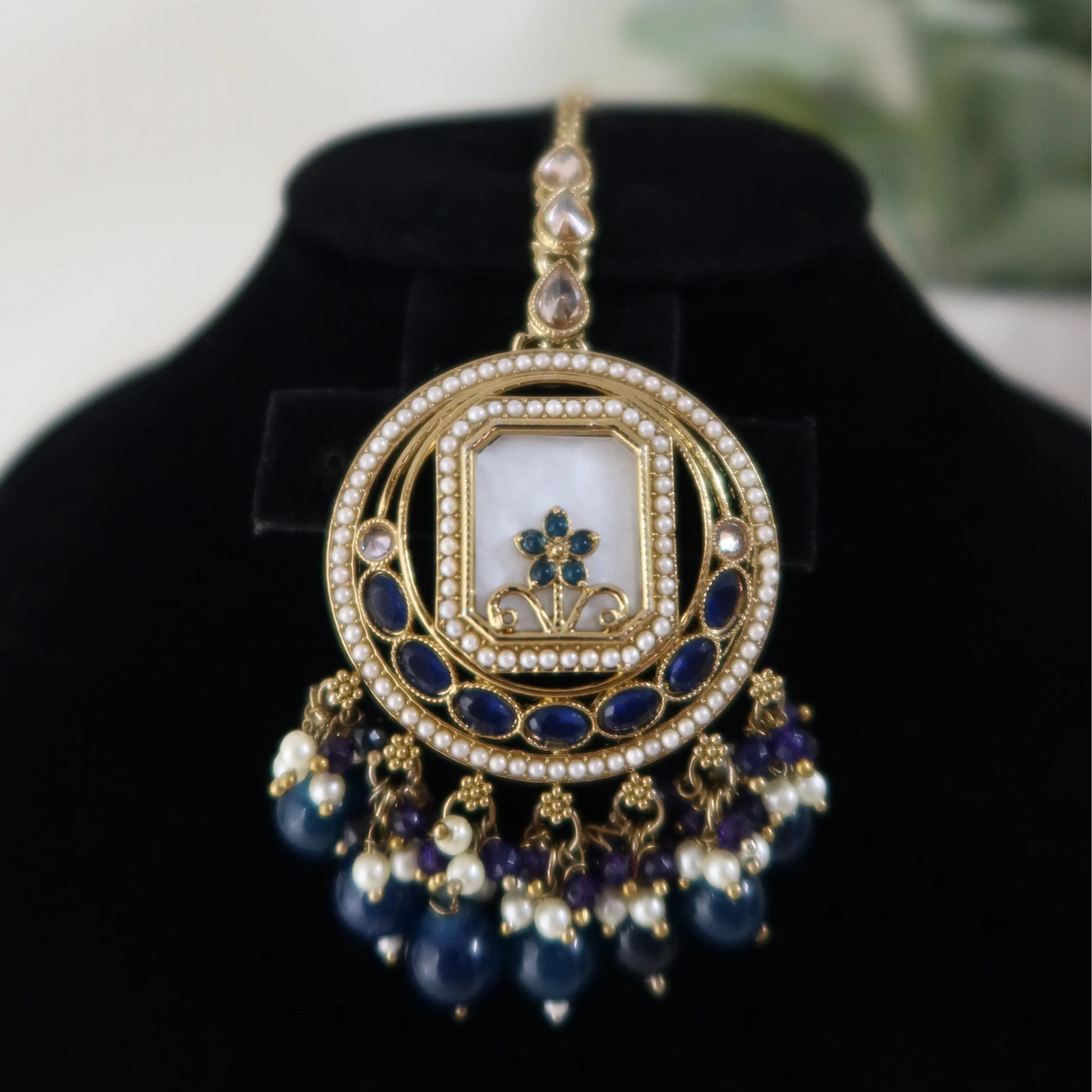Decorative pendant with blue and gold elements on a black stand with a blurred green plant background.