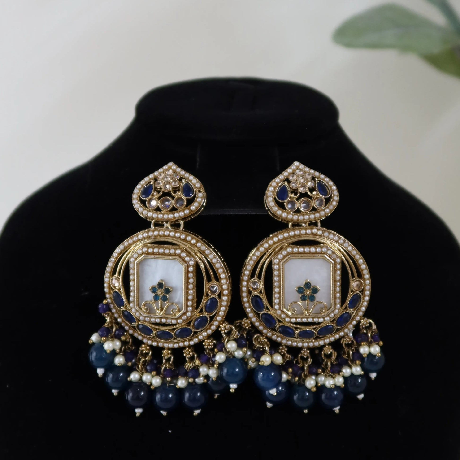 Ornate earrings with blue and gold details on a black stand with a blurred green plant background