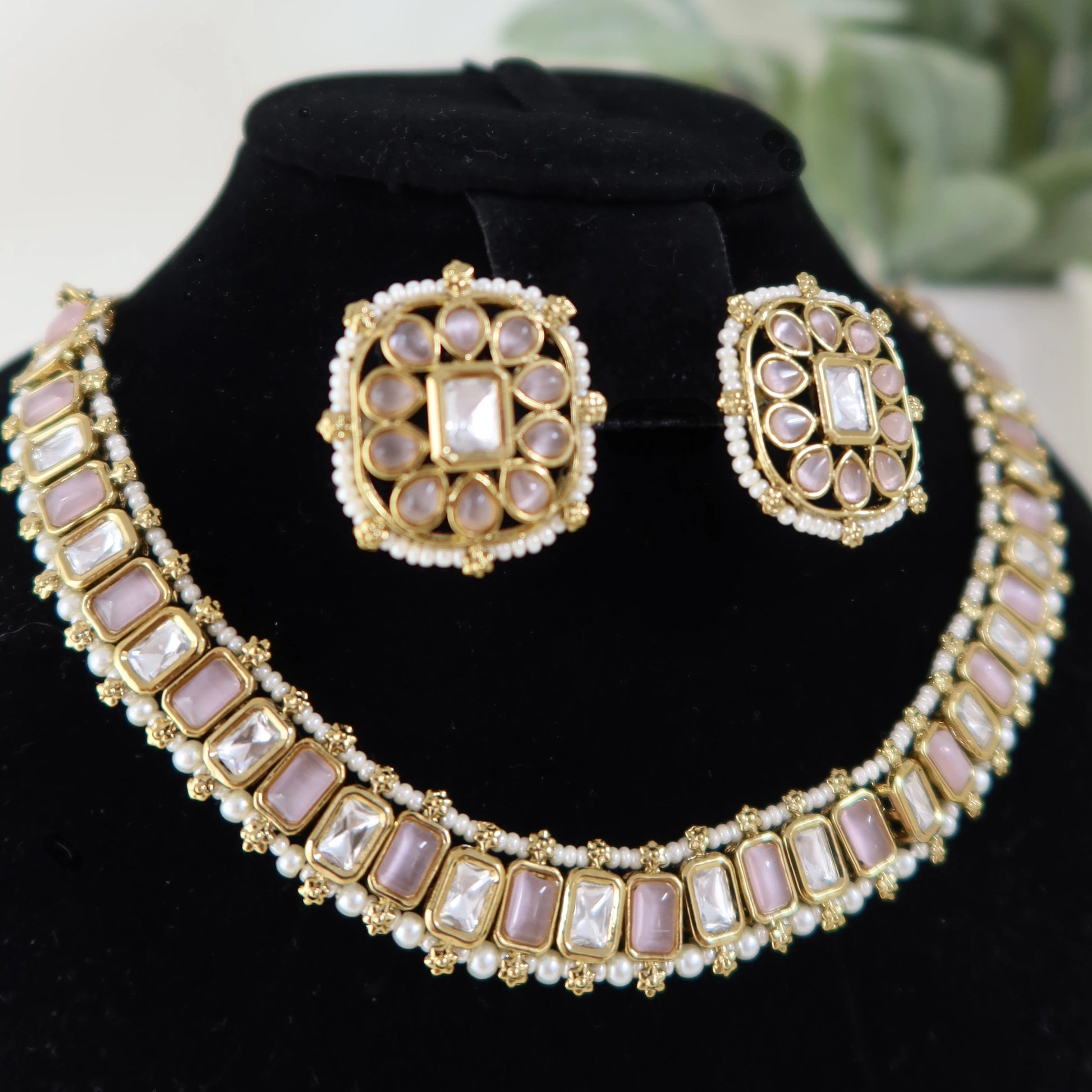 Gold and pearl necklace and earrings set on a black stand with a blurred green plant background