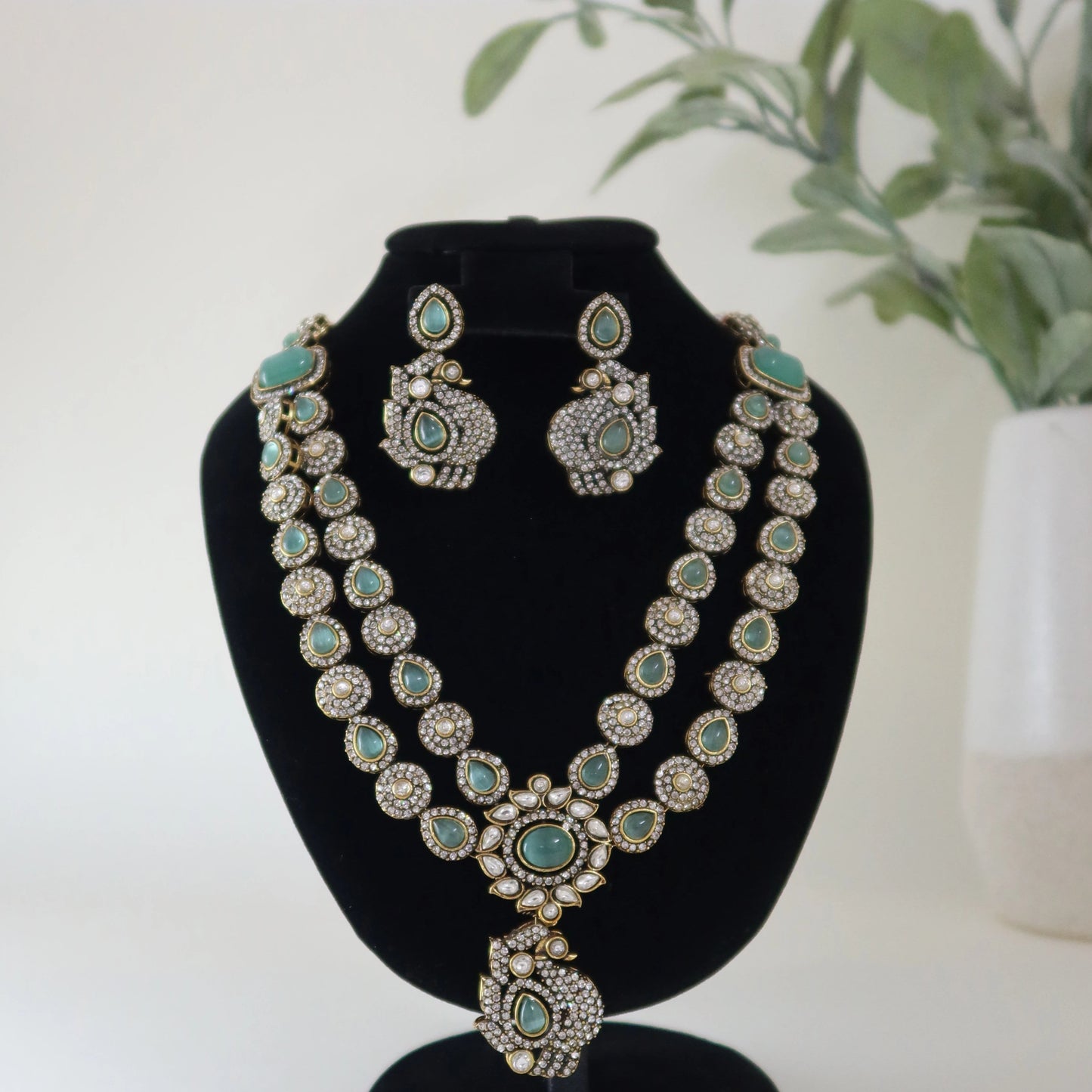Jewelry set with necklace and earrings on a black stand against a white background with a plant.