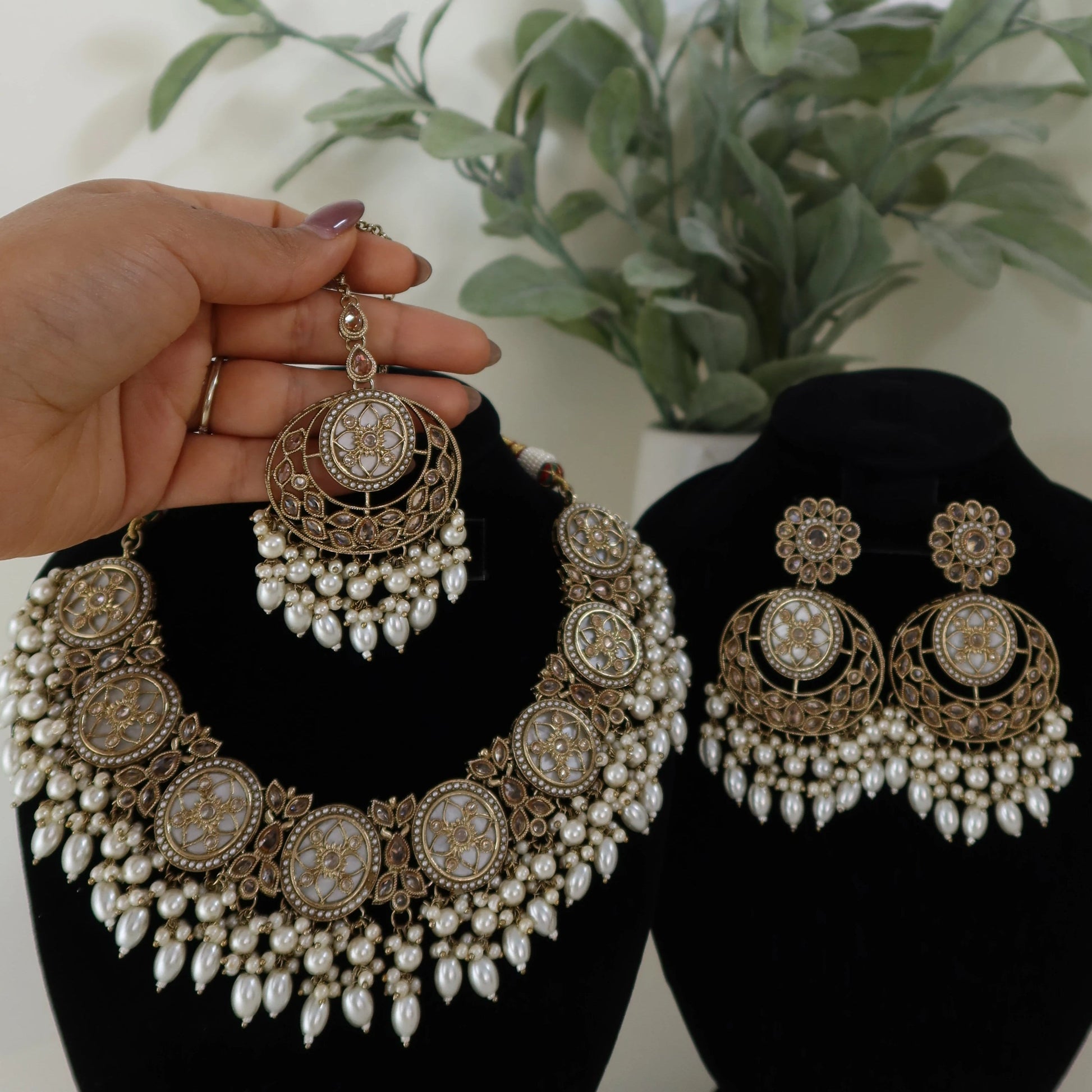 Ornate earrings on black stands with a hand holding one earring, and a plant in the background.