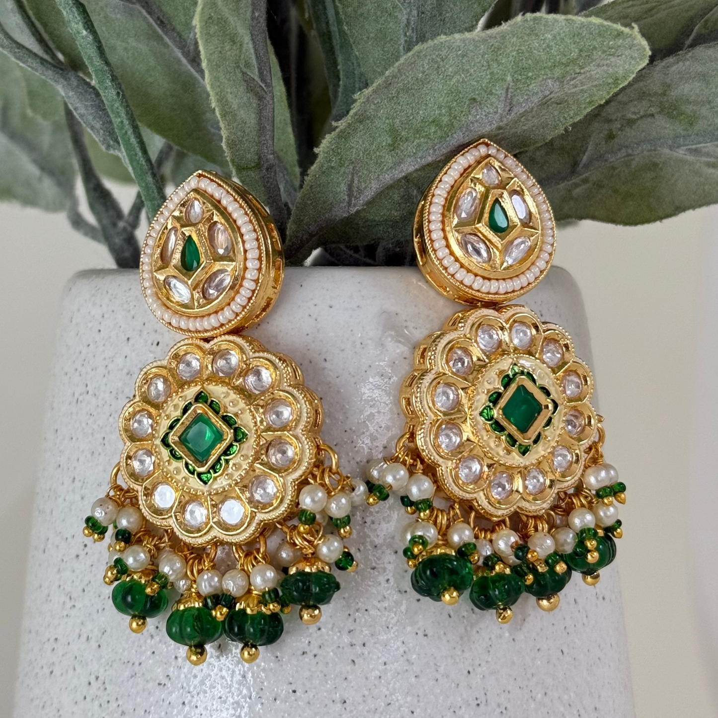 Ornate gold earrings with green and pearl accents on a stone surface with leaves in the background
