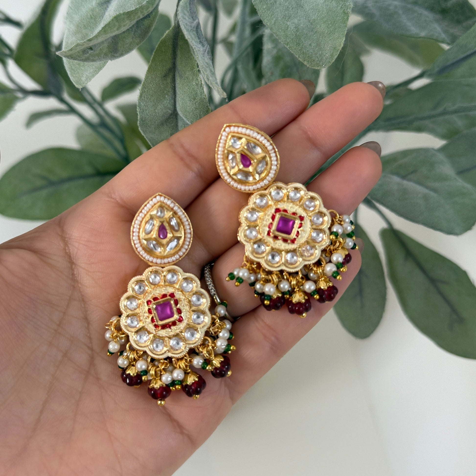 Gold and red earrings held in a hand with a green leafy background