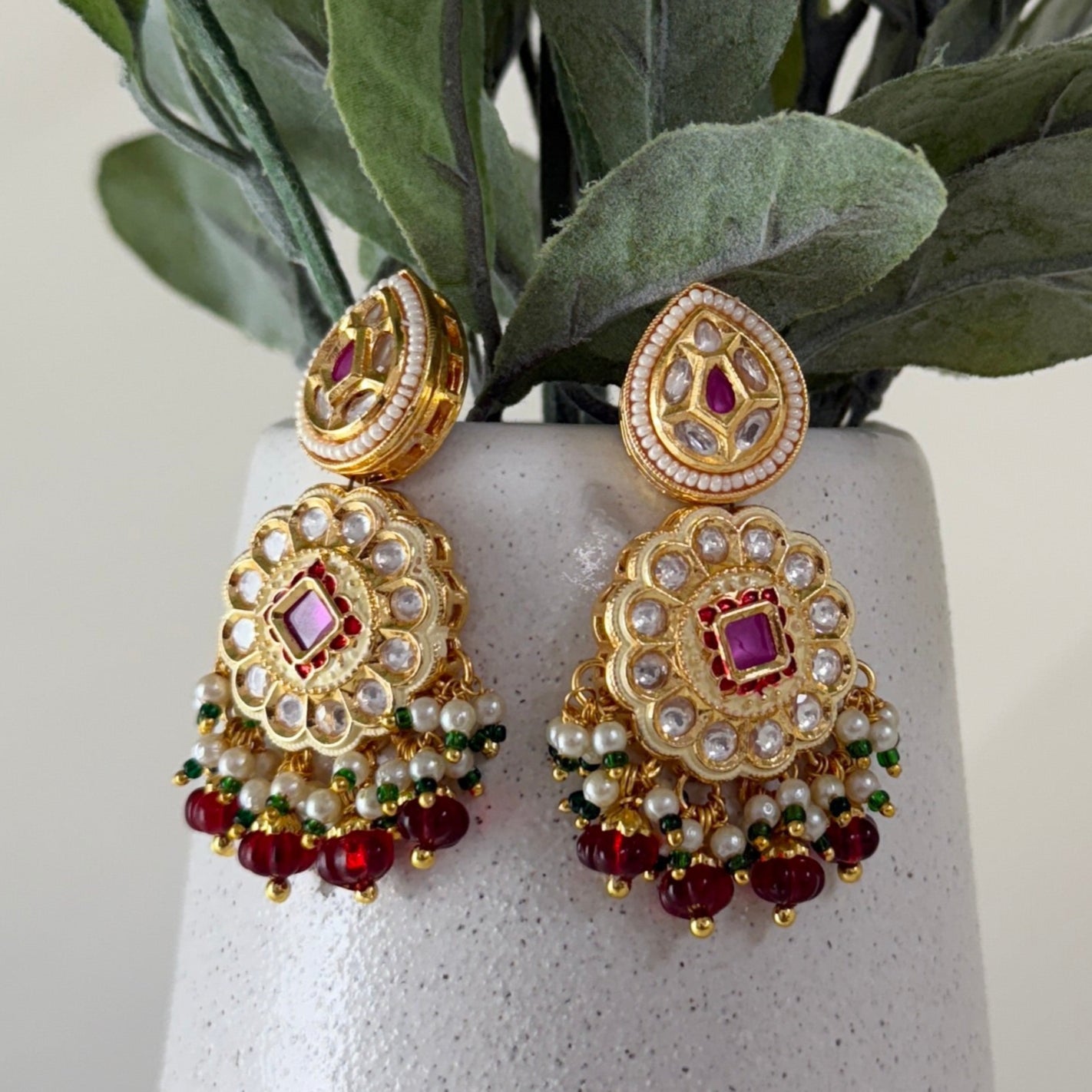 Gold earrings with red and green stones on a white vase with green leaves.