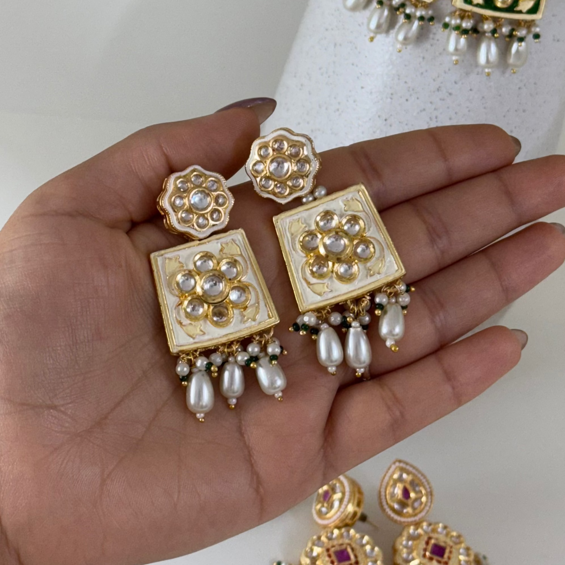 Gold and green earrings held in a hand with a white vase and green leaves in the background.