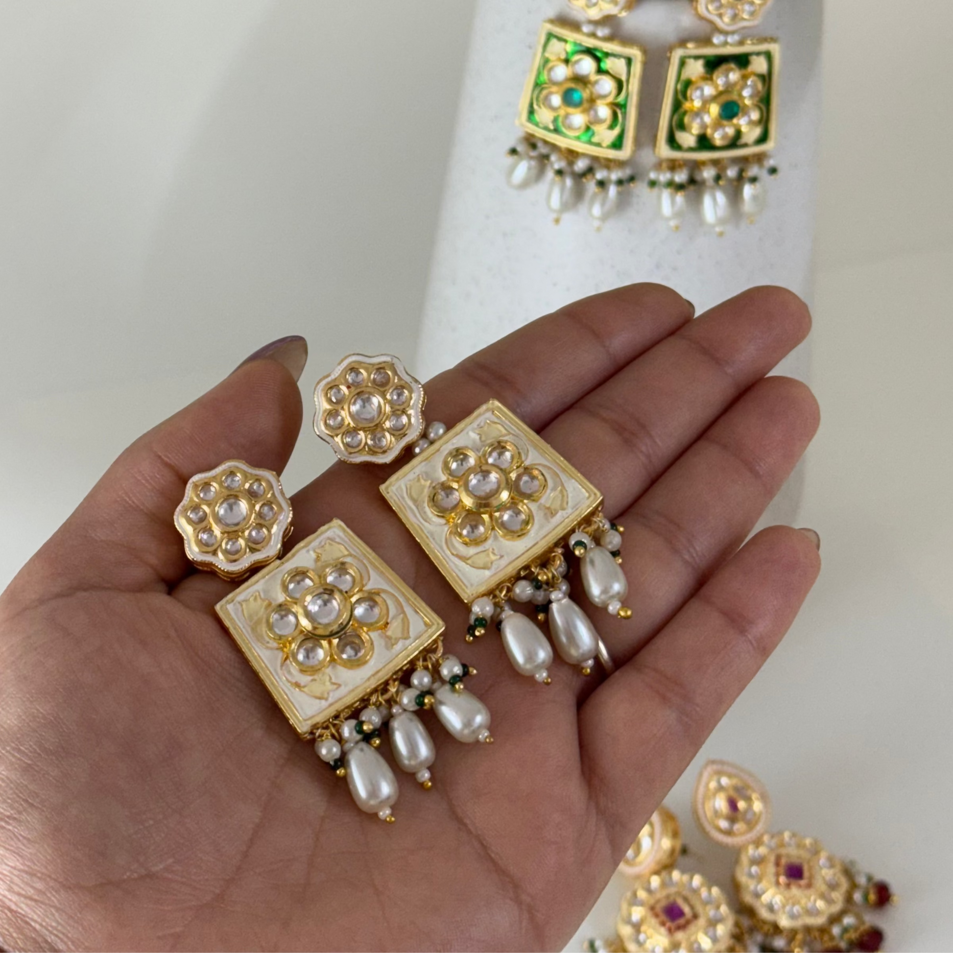 Decorative earrings held in a hand with a white vase and green leaves in the background