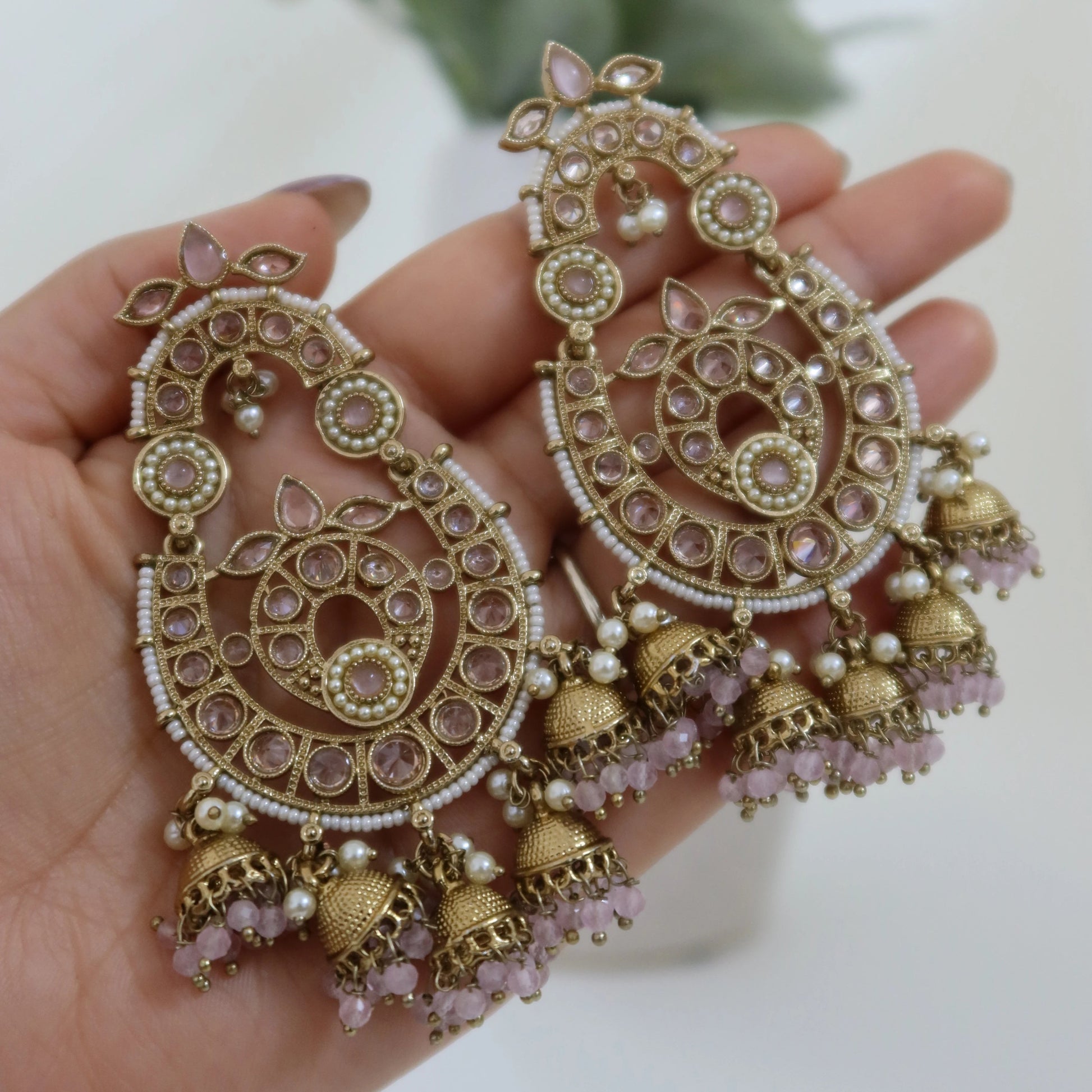 Ornate gold and pink earrings held in a hand with a blurred plant in the background